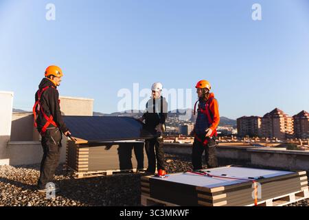 Three workers wearing safety gear are installing solar panels on a rooftop. The image depicts sustainable energy efforts, focusing on clean energy sol Stock Photo
