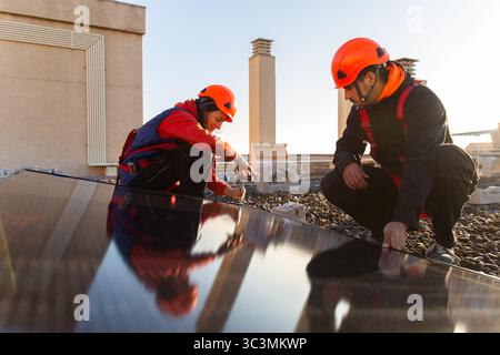 Two workers install solar panels on a building rooftop, wearing safety gear and helmets. The clear sky and urban environment reflect the focus on rene Stock Photo