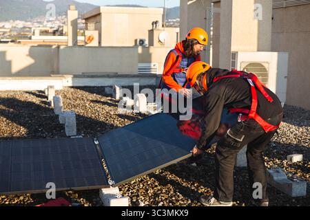 Two skilled workers wearing protective gear install solar panels on a rooftop. The clear sky and urban setting highlight the significance of renewable Stock Photo
