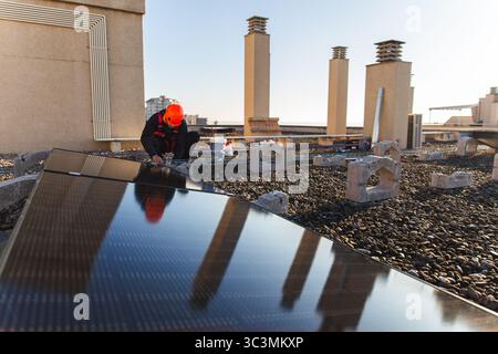 Worker in orange helmet installs solar panels on a city building rooftop. The setup aims to provide renewable energy, harnessing sunlight for sustaina Stock Photo