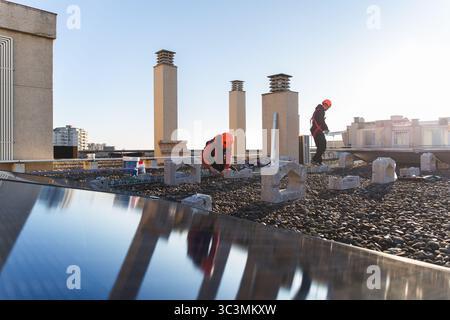 Workers wearing safety gear are installing solar panels on a rooftop. The image captures the modern approach to harnessing solar energy in urban envir Stock Photo
