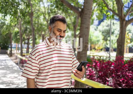Indian man enjoying a summer day in a lush park, smiling at his smartphone, dressed in a striped shirt. The vibrant foliage creates a serene and pleas Stock Photo