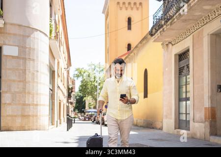 A middle-aged Indian man strolls through sunny city streets with a suitcase in tow. Wearing a light yellow shirt, he checks his phone while enjoying h Stock Photo