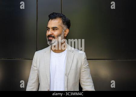 A confident Indian man stands against a modern dark background, wearing a light summer suit and a relaxed expression, embodying the essence of stylish Stock Photo