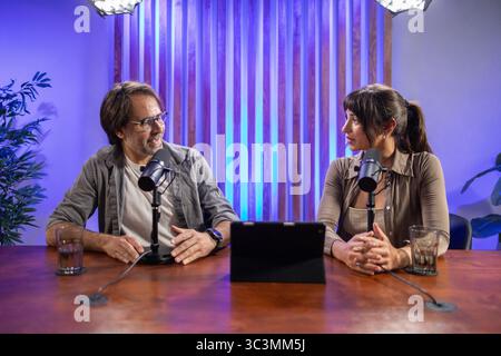 A man and woman sit across from each other at a wooden table, engaging in a lively podcast recording session. Microphones and a digital tablet are vis Stock Photo