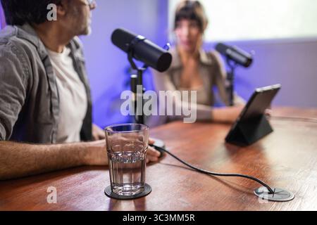 A man and woman are recording a podcast in a modern studio. They are engaged in conversation with professional microphones and a tablet, seated at a w Stock Photo