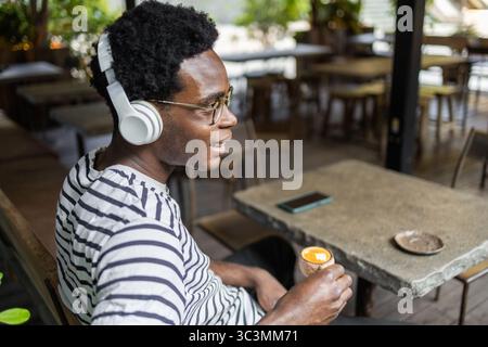 A man sits in a cafe, wearing headphones and holding a coffee. The ambiance is relaxed, with natural light and greenery. A smartphone rests on the tab Stock Photo