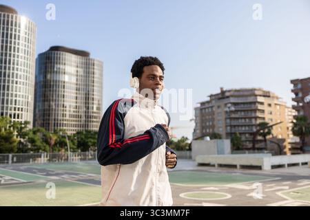 A man jogs confidently in an urban environment, wearing headphones and a tracksuit. Tall buildings line the background, capturing the vibrant city ene Stock Photo