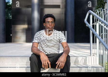 A young male student wearing glasses and a striped shirt sits on outdoor stairs, in front of a contemporary building, appearing focused and thoughtful Stock Photo