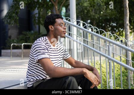 A man with a striped shirt sits on outdoor steps, gazing thoughtfully into the distance, surrounded by greenery and a modern urban environment. Stock Photo
