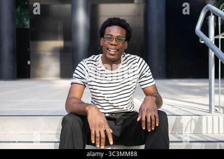A student, dressed casually in a striped shirt and glasses, sits relaxed on campus steps. He exudes positivity and confidence in a modern educational Stock Photo