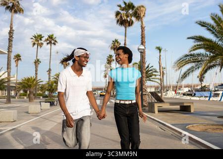 A joyful queer couple walking hand in hand, smiling and laughing under the sun. They're enjoying a summer day surrounded by palm trees and an outdoor Stock Photo