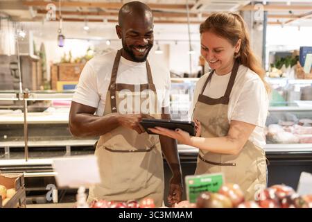 Two cheerful staff members in an eco-friendly store engage over a digital tablet. They are surrounded by fresh produce and sustainable products, promo Stock Photo