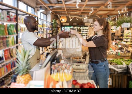 A friendly cashier hands over a reusable bag to a customer at an eco store. Shelves are filled with organic products, promoting sustainability and a g Stock Photo