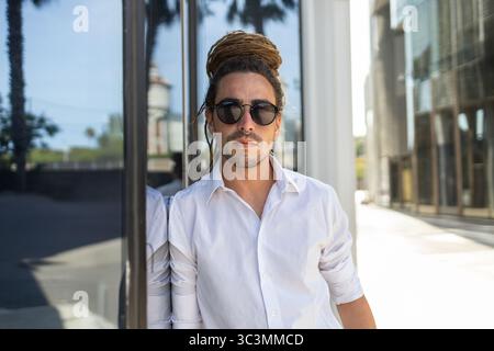 An Italian man with dreadlocks stands confidently against a building during his travels in Spain. He wears a white shirt and sunglasses, exuding a sty Stock Photo