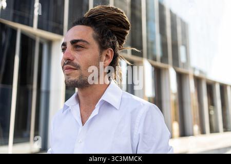 Young Italian man with dreadlocks enjoying travel in Spain. He stands confidently in front of modern architecture, embracing a city vibe with a sense Stock Photo