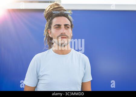 A stylish Italian man with dreadlocks stands confidently against a blue backdrop, enjoying his travel experience in sunny Spain. He exudes a carefree, Stock Photo