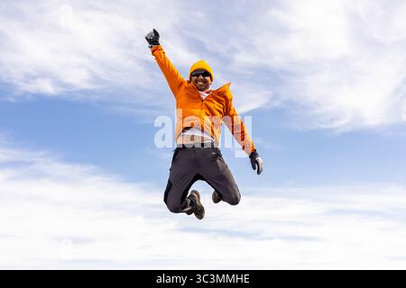 A man leaps joyfully in a breathtaking mountain landscape, wearing an orange jacket and yellow cap. The adventure captures the essence of freedom and Stock Photo