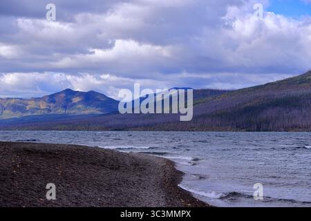 A sweeping view of Lake McDonald’s stony shoreline and rippling waters under dramatic clouds, with forested mountains leading toward a serene horizon. Stock Photo