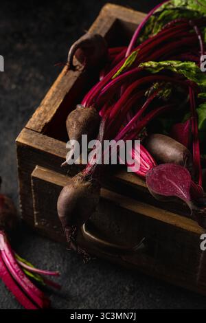Fresh beetroots with vibrant green stems and leaves are displayed in a rustic wooden box on a dark surface. The rich colors and textures make a striki Stock Photo