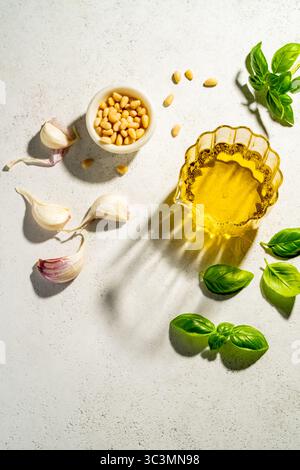 Top view of basil leaves, a glass bowl of olive oil, garlic cloves, and pine nuts on a textured white background, showcasing fresh ingredients for mak Stock Photo