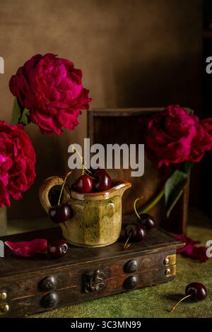 A rustic scene showcasing vibrant red cherries in a ceramic jug, surrounded by lush pink peonies. The setting rests on a textured wooden surface with Stock Photo