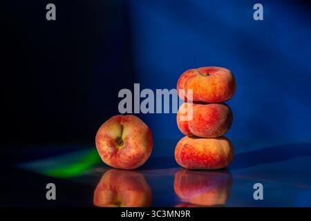 A stack of three flat peaches and one beside it sit on a reflective surface. The dark blue background enhances their vibrant colors and textures, crea Stock Photo