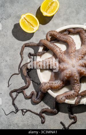 A fresh octopus elegantly arranged on a white plate, accompanied by two lemon halves on a textured gray surface, perfect for a seafood themed presenta Stock Photo