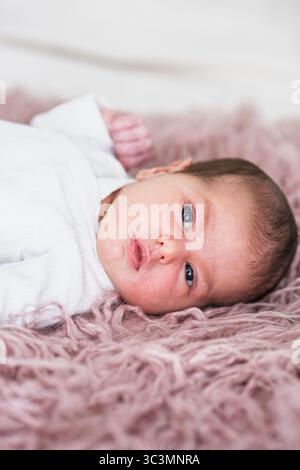 A newborn baby peacefully lying on a soft pink rug, wrapped in a cozy white onesie. The gentle texture and serene ambiance create a warm, comforting a Stock Photo