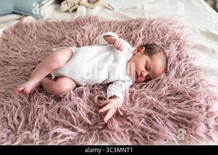 A serene newborn baby comfortably lies on a soft, fluffy pink blanket, dressed in a white onesie. The background suggests a cozy, warm, and peaceful s Stock Photo