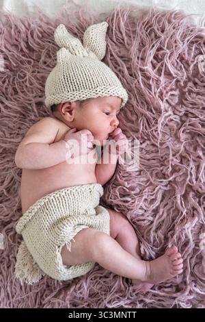 Newborn baby resting peacefully on a soft, fluffy pink blanket, wearing a cozy knitted hat with bunny ears. The warm tones add to the gentle and seren Stock Photo