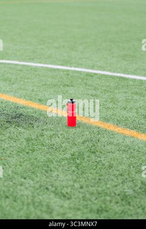 A vibrant red water bottle rests on a soccer field's artificial green turf, surrounded by painted white and orange lines. Ideal for depicting sports c Stock Photo