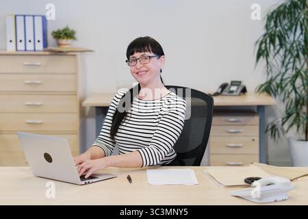 Secretary smiles at camera, works on laptop Stock Photo