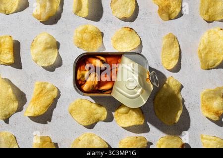 Top view of an open can of mussels surrounded by scattered potato chips on a textured stone background. The composition highlights the contrast betwee Stock Photo