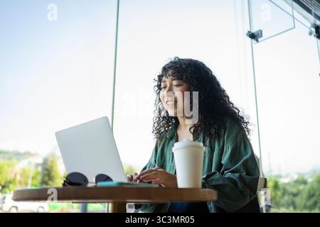 A curly-haired young woman enjoys working on her laptop in a bright, modern cafe. She sits at a wooden table, surrounded by natural light and a citysc Stock Photo