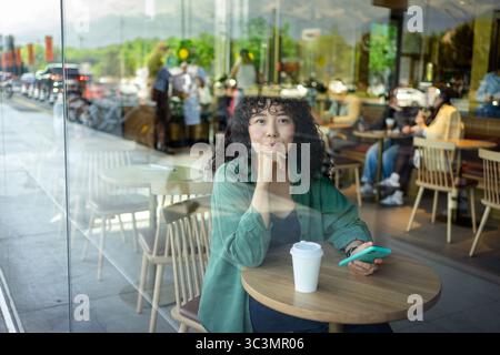 A young woman with curly hair sits at a cafe table, holding a smartphone and enjoying a coffee. The atmosphere is relaxed with people in the backgroun Stock Photo