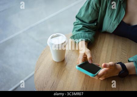 A woman relaxes at a cafe table, holding a smartphone in one hand, with a takeaway coffee cup nearby. She wears a green jacket and a black watch, enjo Stock Photo