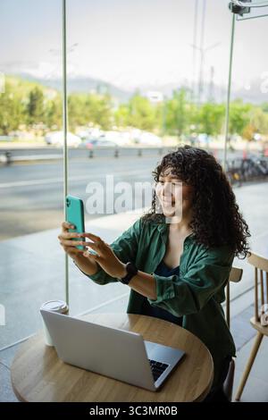 A young woman with curly hair sits at a cafe table, smiling as she takes a selfie with her smartphone. A laptop and coffee cup are on the table, windo Stock Photo