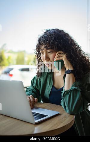 Curly haired young woman sits at a wooden table working on a laptop while talking on the phone. Natural light from the window illuminates her focused Stock Photo