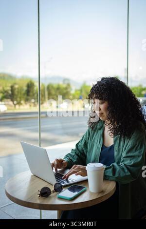 A young woman with curly hair is working on her laptop at a cafe. She looks focused and comfortable in a casual green shirt, with a coffee cup and sma Stock Photo