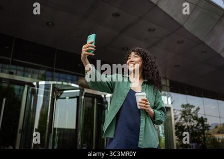 A cheerful, curly-haired young woman takes a selfie outside a modern building, holding a coffee cup. She's dressed casually and enjoys the sunny day i Stock Photo