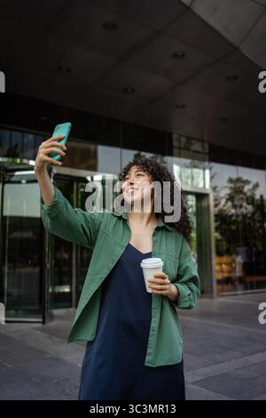 Curly-haired young woman stands outside a modern building, smiling as she takes a selfie with her smartphone. She holds a takeaway coffee cup, capturi Stock Photo