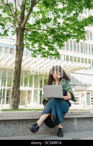 Curly-haired young woman works on her laptop while sitting outdoors in a city environment, enjoying the fresh air and natural surroundings near an urb Stock Photo