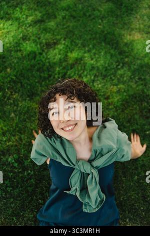 A young woman with curly hair is sitting on the grass, smiling brightly at the camera. She looks relaxed and content, with a green scarf draped around Stock Photo