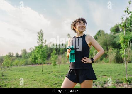 A young woman with curly hair is smiling as she holds a water bottle. She stands confidently in a green field under a clear sky, ready for an outdoor Stock Photo