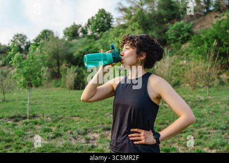 A young woman with curly hair drinks from a water bottle while enjoying a refreshing outdoor workout. She stands in a lush, green park, emphasizing he Stock Photo
