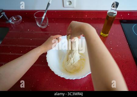 A woman skillfully kneads dough in a white bowl on a vibrant red countertop, preparing homemade focaccia using fresh ingredients and olive oil in a mo Stock Photo