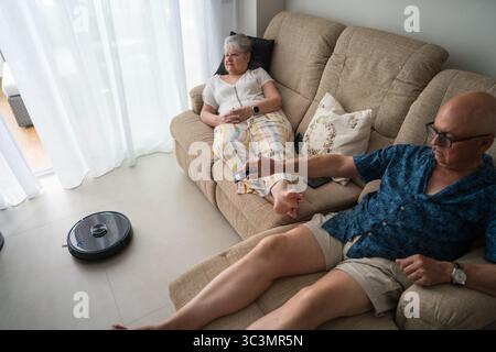 A senior couple relaxes at home while using a smart robot vacuum, embodying vitality and independence with modern technology in their everyday digital Stock Photo