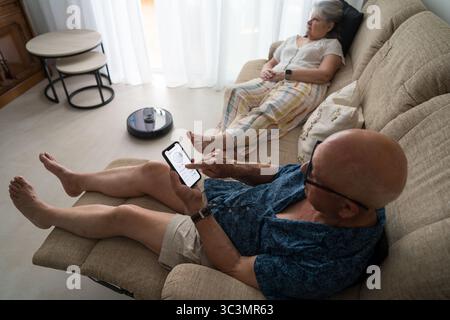 A senior couple relaxes at home while using a smart robot vacuum. This image captures the integration of smart home technology and active senior livin Stock Photo