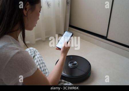 An Asian woman sits on the floor, using a smartphone app to control a robotic vacuum cleaner. The clean, minimalist setting highlights modern technolo Stock Photo
